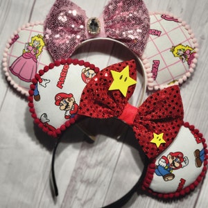 Puede incluir: Dos conjuntos de orejas de ratón temáticas. Un conjunto presenta lazos de lentejuelas rosas y orejas de la Princesa Peach. El otro conjunto tiene lazos de purpurina roja con detalles de estrellas amarillas y orejas de Mario. Ambos conjuntos tienen diademas negras.