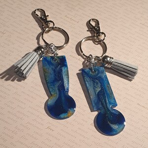 Op de afbeelding: Twee sleutelhangers van hars met blauwe en gouden marmerpatronen. Elke sleutelhanger heeft een zilveren sleutelring en een grijze kwast.