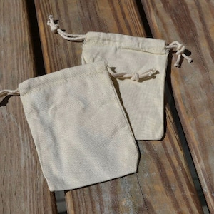 May include: Two small, white cotton drawstring bags on a wooden surface. The bags are empty and have a natural, unbleached look.