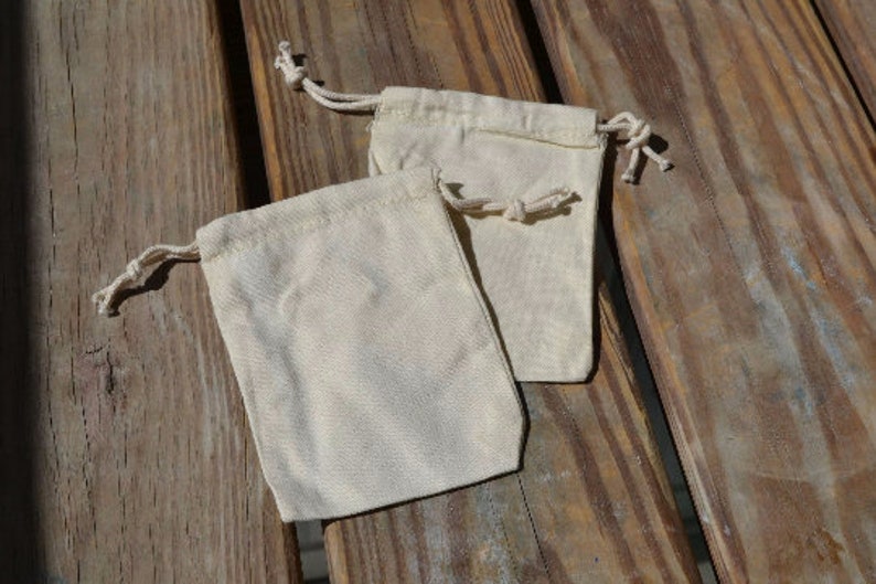 May include: Two small, white cotton drawstring bags on a wooden surface. The bags are empty and have a natural, unbleached look.