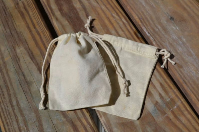 May include: Two white cotton drawstring pouches with a natural cotton cord. The pouches are lying on a wooden surface.