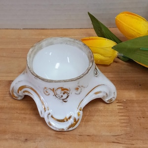 May include: White ceramic egg holder with a gold trim and a decorative base with three legs. The holder is on a wooden surface.