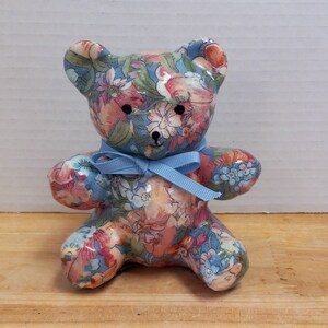 May include: A small, ceramic teddy bear figurine with a floral pattern in shades of blue, pink, and green. The bear is wearing a blue ribbon around its neck.