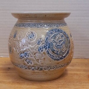 May include: A small, round ceramic vase with a beige glaze and blue and white floral designs. The vase has a slightly textured surface and a wide opening.