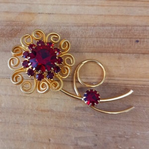 May include: A gold brooch with a red rhinestone flower design. The flower has a large center stone surrounded by smaller stones. The brooch has a delicate stem with a small red rhinestone at the end.