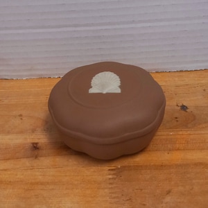 May include: A brown, round trinket box with a white seashell design on the lid. The box is sitting on a wooden surface. The box has a smooth, matte finish and a scalloped edge.