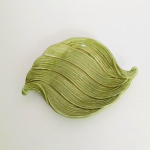May include: A light green, leaf-shaped ceramic dish. The dish has a textured surface with a series of curved lines, mimicking the veins of a leaf. The edges of the dish are gently curved, and the overall shape is organic and natural.