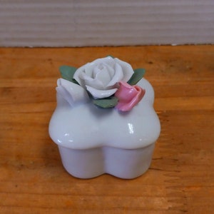May include: A white ceramic trinket box with a flower design. The box is shaped like a four-leaf clover and has a white rose with pink accents.