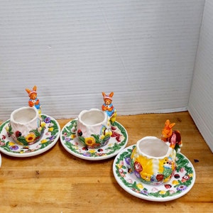 May include: Four ceramic teacups with bunny rabbit handles and floral designs. Each cup is on a matching saucer with a green, yellow, and red floral pattern.