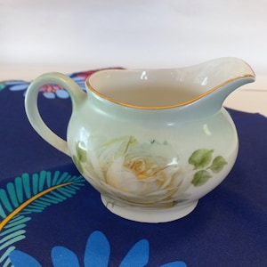 May include: A white porcelain creamer with a delicate floral design featuring a white rose with green leaves. The creamer has a gold rim and is sitting on a blue and white patterned fabric.