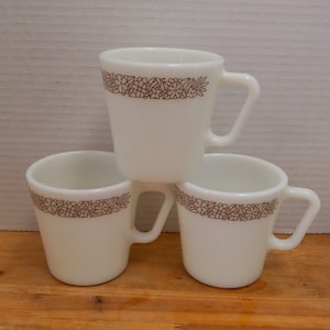 May include: Three white ceramic mugs with brown floral designs around the top. The mugs are stacked on top of each other.