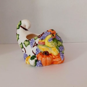 May include: A white ceramic cornucopia with a brown stem and leaves, filled with colorful fall fruits and vegetables, including grapes, pumpkins, and gourds.