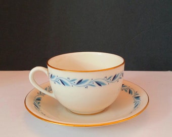 May include: A white teacup and saucer with a blue and gold floral pattern. The cup has a rounded shape and a small handle. The saucer has a scalloped edge.