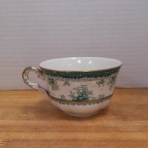 May include: A white teacup with a green and gold floral pattern. The cup has a handle and is sitting on a wooden surface.