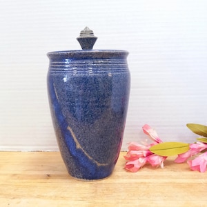 May include: A blue ceramic jar with a lid. The jar has a speckled glaze and a light blue stripe around the top. The lid is a simple, pointed shape.
