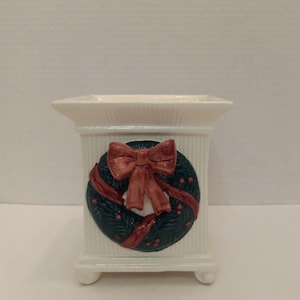 May include: White ceramic planter with a raised green and red Christmas wreath design. The wreath has a red bow and red berries.