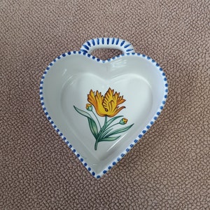 May include: A white heart-shaped ceramic dish with a blue and white trim. The dish features a yellow flower with green leaves painted on the inside.