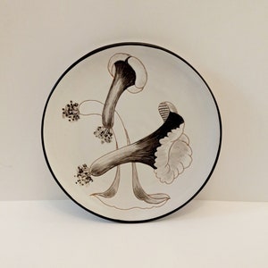 May include: White ceramic plate with a black rim featuring a stylized design of two brown and black flowers.