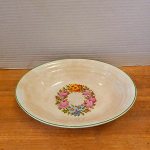 May include: A white ceramic bowl with a green rim and a floral design in the center. The design features a wreath of pink, blue, and yellow flowers.