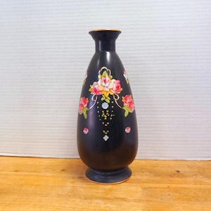 May include: A black vase with a floral design. The vase has a narrow neck and a wider base. The floral design features pink and white roses with green leaves. The rim of the vase is gold. The vase is on a wooden surface.
