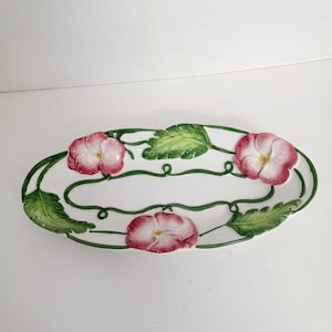 May include: Oval ceramic serving dish with a white base, adorned with pink flower and green leaf designs. The flowers have yellow centers. The rim is decorated with green vines and leaves.