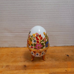 May include: A white porcelain egg-shaped trinket box with a floral design in shades of pink, yellow, blue, and red. The box has a gold trim and sits on three gold feet.