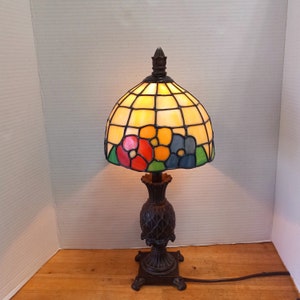May include: A stained glass table lamp with a floral design. The lampshade is made of clear glass with colorful flowers and leaves. The base is a dark brown metal with a pineapple-shaped design.