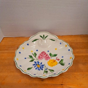 May include: A white ceramic shell-shaped dish with a green, yellow, pink, and blue floral design. The dish has a raised rim and a scalloped edge.
