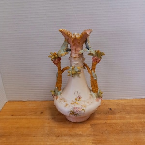 May include: A white porcelain vase with a pink and gold floral design. The vase has three handles that are shaped like stylized palm trees. The vase is decorated with painted flowers and leaves.