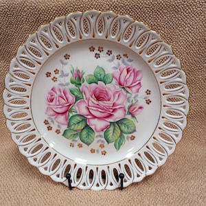 Puede incluir: Plato de porcelana blanca con borde dorado y un diseño de corte complejo. El centro presenta un diseño floral pintado a mano de rosas rosas con hojas verdes y detalles dorados.