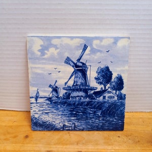 May include: A blue and white ceramic tile depicting a Dutch windmill scene. The tile features a windmill with a white body and blue blades, a smaller windmill in the background, a small house, and a river.