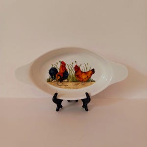 May include: White oval ceramic baking dish with a rooster and hen design. The rooster is black and red, and the hen is brown and red. The dish has two handles.