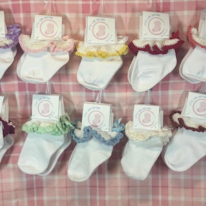 Könnte beinhalten: Eine Sammlung weißer Babysocken mit farbigen gehäkelten Spitzenborten in verschiedenen Farben, darunter Lila, Rosa, Gelb, Rot, Grün und Blau. Jedes Paar wird mit einem kleinen Etikett präsentiert. Die Socken werden vor einem rosa-weiß karierten Hintergrund ausgestellt.