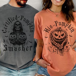 Può includere: Due magliette a tema Halloween. La prima maglietta è grigia con il testo "Certified Pumpkin Smasher" e uno scheletro che tiene due zucche. La seconda maglietta è arancione con il testo "His Pumpkin" e una zucca con un teschio.