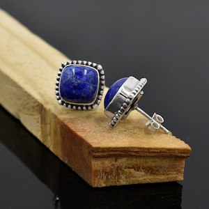 May include: A pair of silver stud earrings with square blue lapis lazuli gemstones. The earrings have a beaded border around the gemstones.