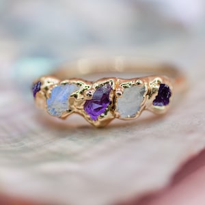 May include: A gold ring set with five raw gemstones. The ring features two amethyst stones and three moonstone stones.