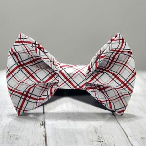 May include: A white bow tie with a red, black, and gray plaid pattern. The bow tie is centered on a white wooden surface. The bow tie is a classic accessory.