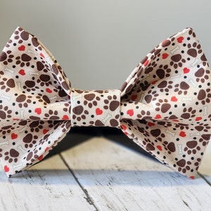 Könnte beinhalten: Eine beige Schleife mit einem sich wiederholenden Muster aus braunen Pfotenabdrücken, roten Herzen und weißen Knochen. Die Schleife ist in der Mitte gebunden und für Haustiere bestimmt.