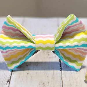 May include: A colourful bow tie with a wavy striped pattern in shades of pink, yellow, green, and blue. The bow tie is made of fabric and is tied in a classic bow shape.