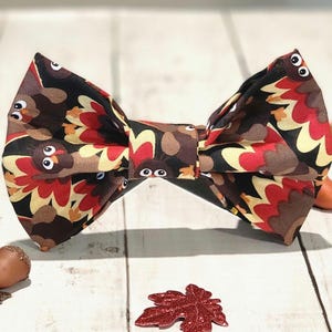 May include: A brown, red, and yellow bow tie with a turkey print. The bow tie features a repeating pattern of turkeys with brown feathers and red wattles.