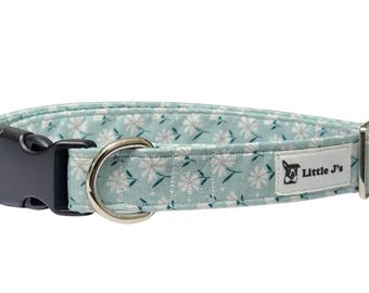 May include: A light blue dog collar with a white daisy floral pattern. The collar has a black plastic buckle, a silver metal D-ring, and a label that reads "Little J's".