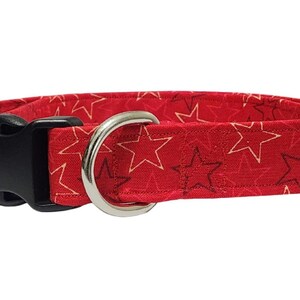 May include: Red dog collar with a star pattern. The collar has a black plastic buckle, a silver metal D-ring, and a silver metal slide. The fabric features a repeating pattern of white and dark red stars.