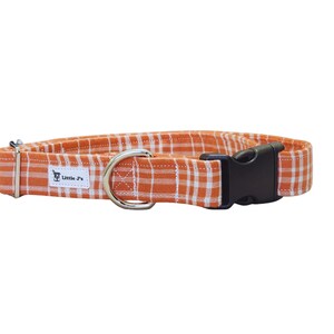 May include: An orange and white plaid fabric pet collar featuring a black plastic side-release buckle, a silver metal D-ring for lead attachment, and a small white rectangular fabric label with the text Little J's printed in black.