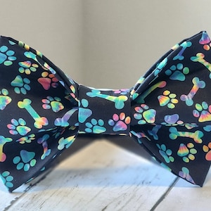 May include: A black bow tie with a colorful paw print and bone pattern. The paw prints are tie-dye style and the bones are white.