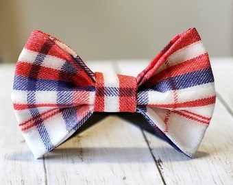 May include: A red, white, and blue plaid bow tie. The bow tie features a classic butterfly shape with a repeating pattern of red, white, and blue squares and stripes. The fabric appears to be a woven material.