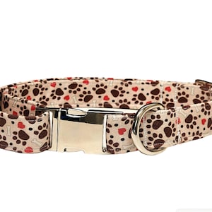 May include: A beige dog collar with a paw print and heart pattern. The collar has a silver buckle and a silver D-ring.