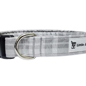May include: A grey and white plaid dog collar with a black plastic buckle and a silver metal D-ring. The collar features a white label with the text "Little J's" and a dog logo.