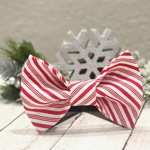 May include: A red and white striped bow tie with a candy cane pattern. The bow tie is made of fabric and is tied in a classic bow shape.