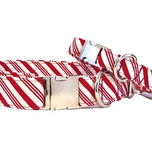 May include: Two dog collars with a red and white candy cane stripe pattern. The collars have silver metal hardware, including buckles and D-rings. One collar is larger than the other, suggesting different sizes.