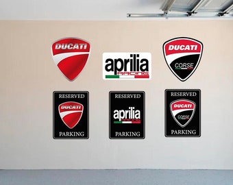Puede incluir: Tres señales de estacionamiento reservado con los logotipos de Ducati Corse, Aprilia Racing y Ducati. Las señales son negras con texto blanco y están montadas en una pared beige.
